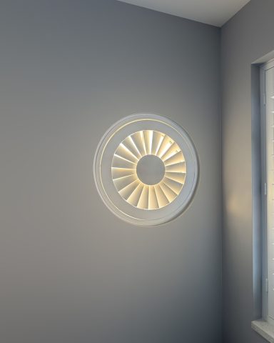 Round window with slatted design, casting light onto a grey wall corner.